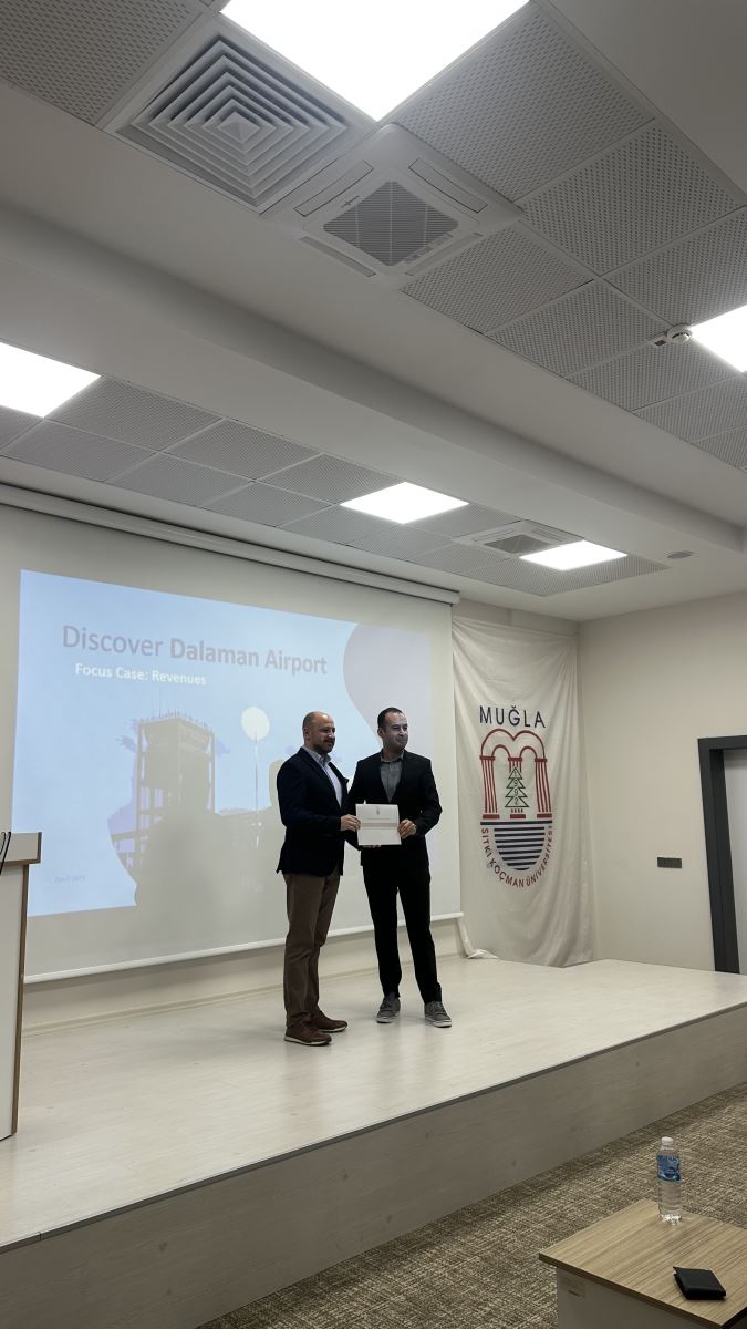 Ahmet Orçun Songur and a faculty member are standing side by side in the center of the stage. Both are smiling and holding a certificate of appreciation together. In the background, the screen displays a slide titled "Discover Dalaman Airport," and the emblem of Muğla Sıtkı Koçman University is visible on the right.
